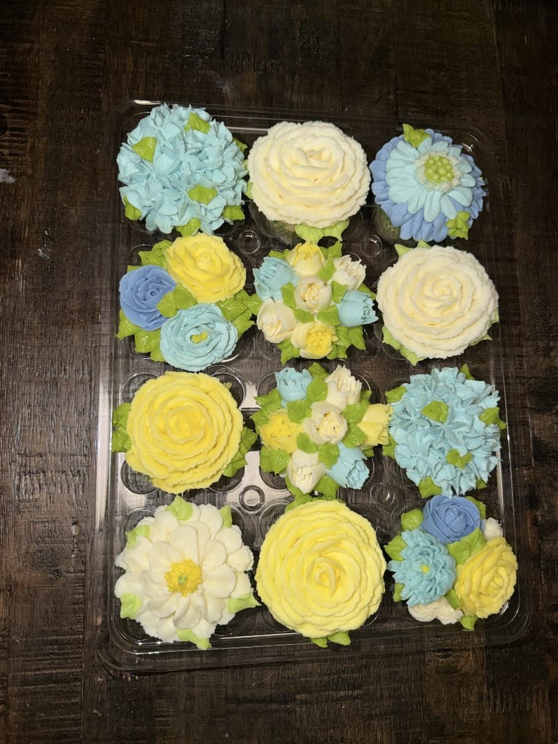 Twelve buttercream flower cupcakes in blue, yellow, and white on a dark wood table