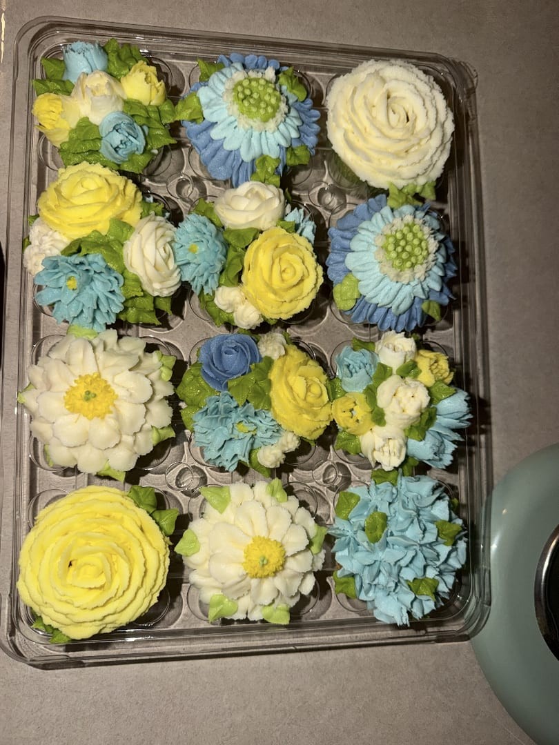 A tray of a dozen floral cupcakes piped in yellow, blue, and white buttercream