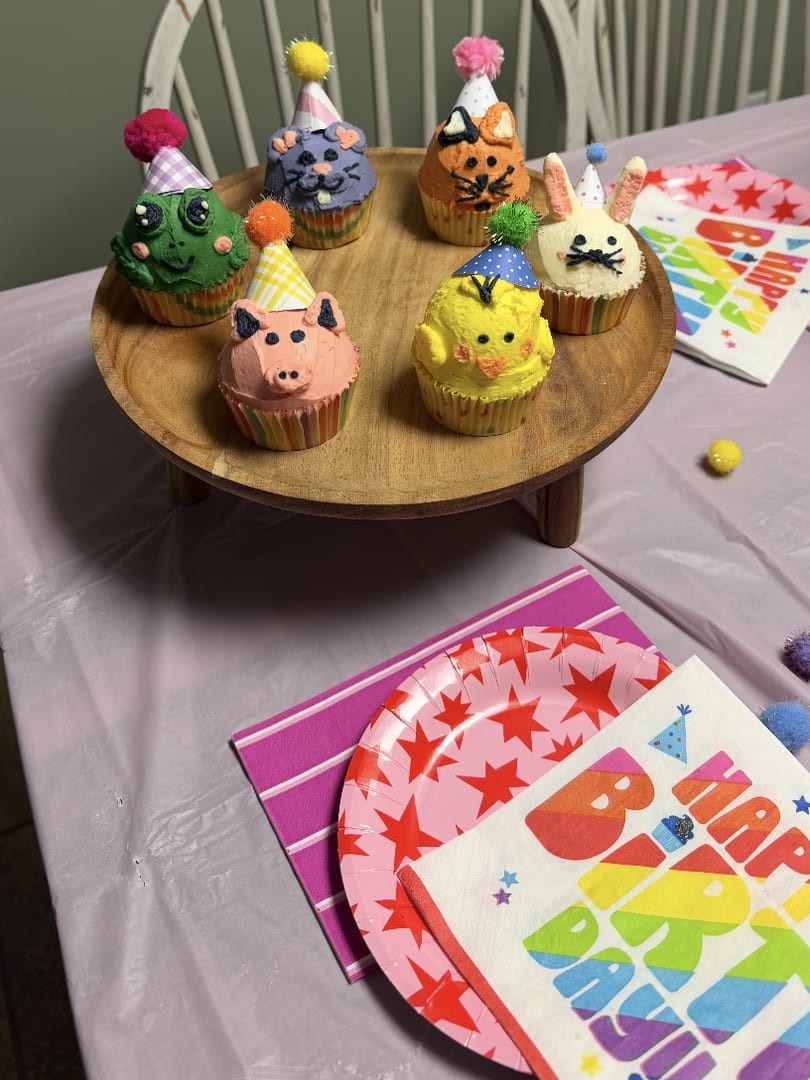 Animal-face cupcakes wearing party hats on a wooden cake stand at a birthday table