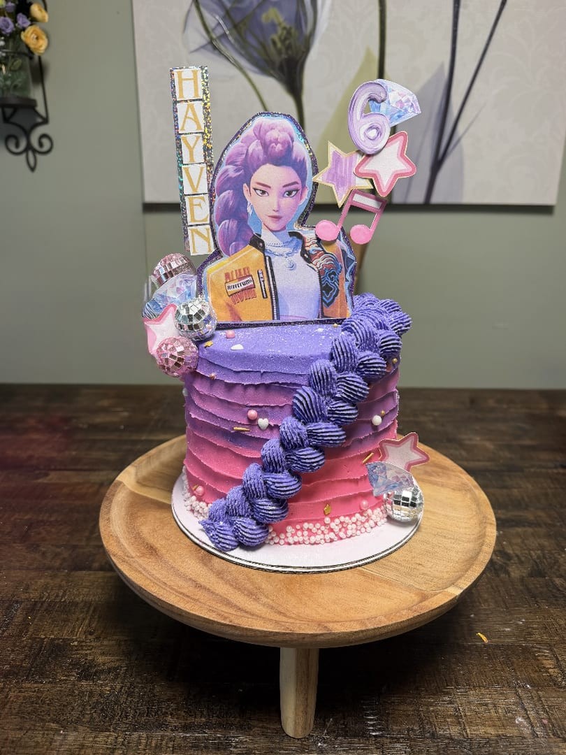 Tall purple and pink ombre cake with a character cutout topper on a wooden stand