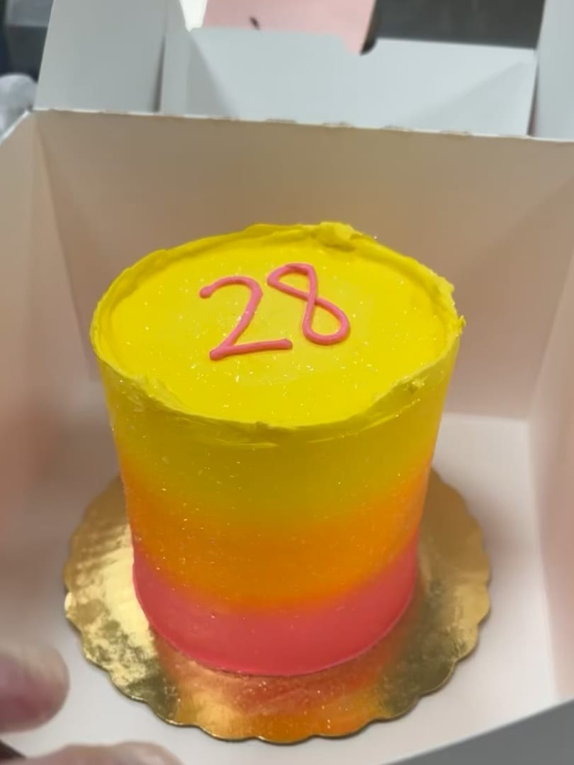 Small tall cake with yellow-to-pink ombre frosting and a red 28, in a cake box