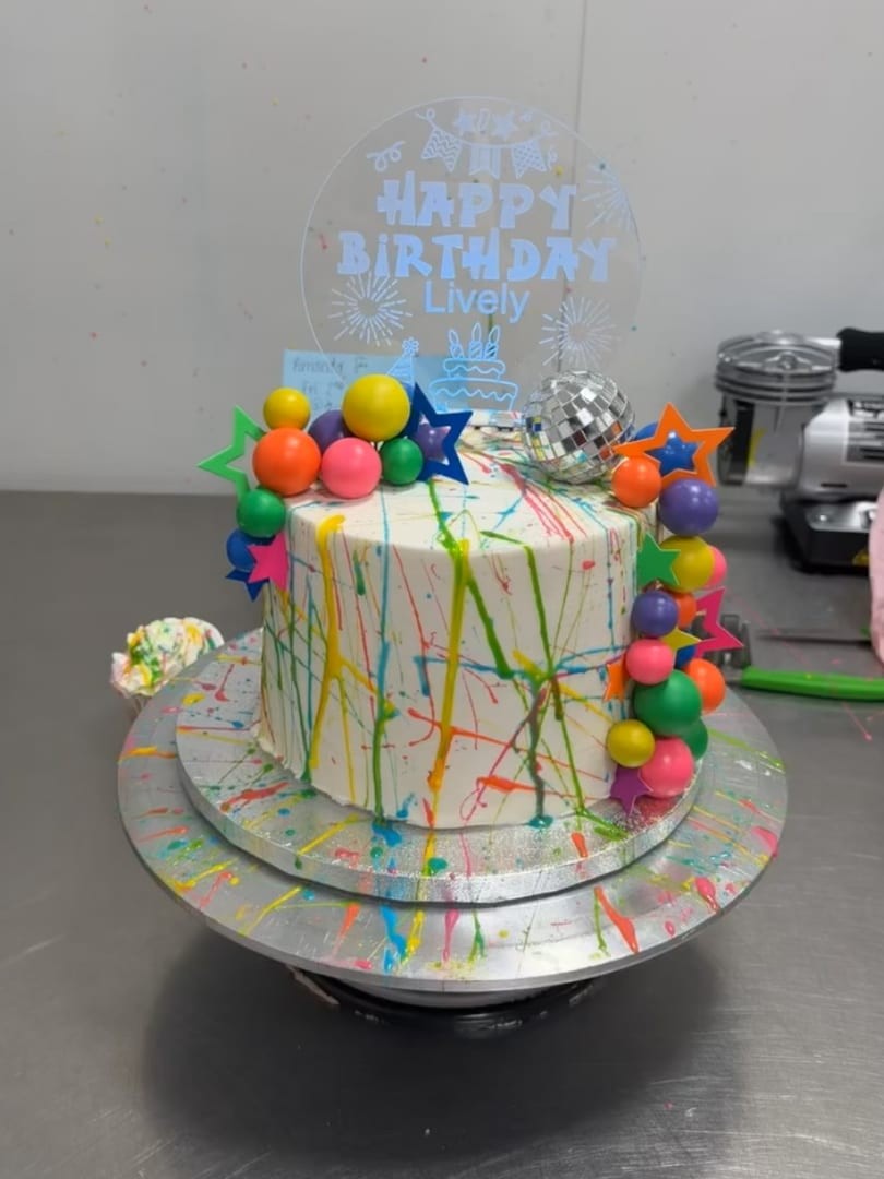 Tall white cake with paint-splatter drips, bright balloons, and a clear birthday topper